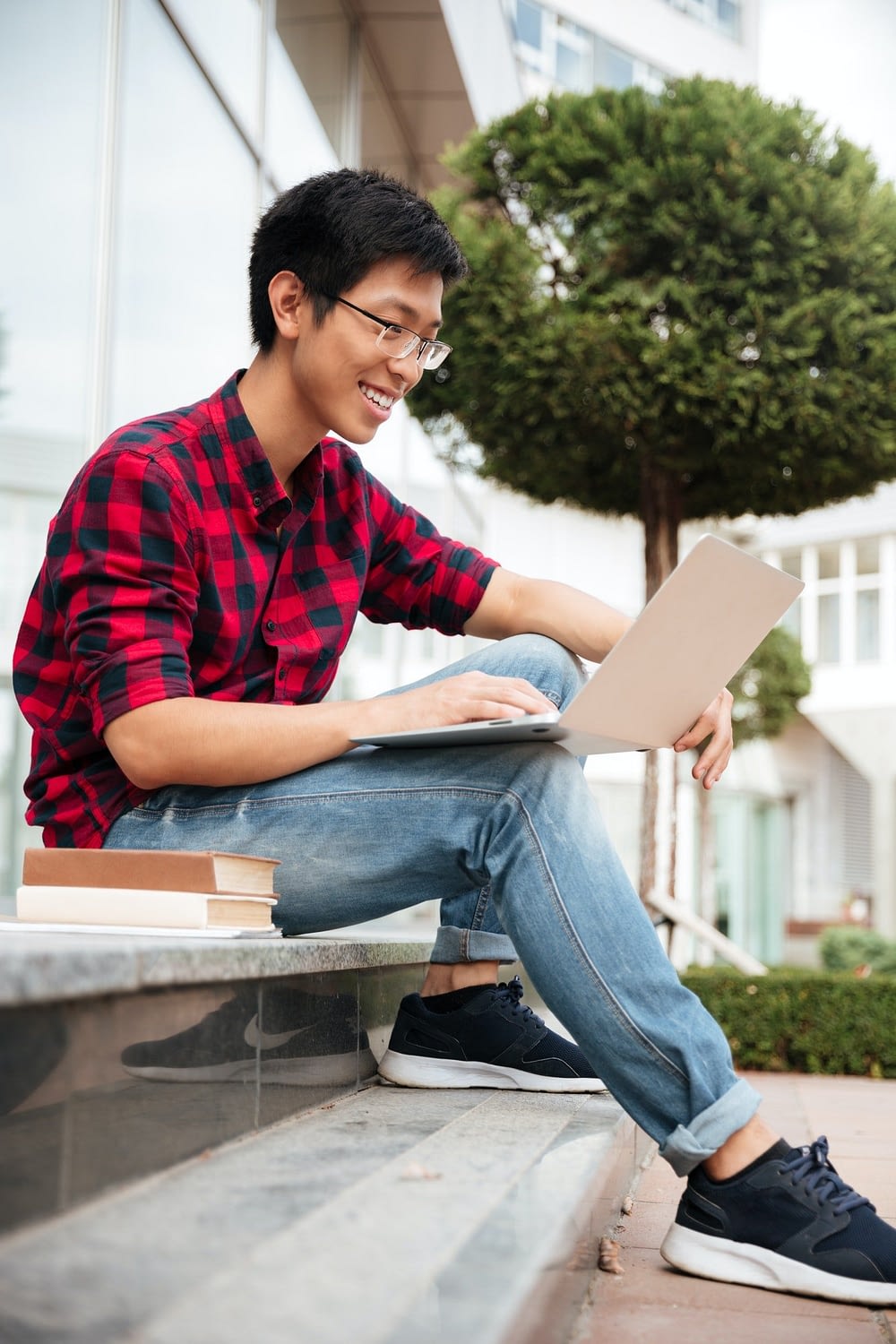 Cheerful asian young man sitting and using laptop outdoors